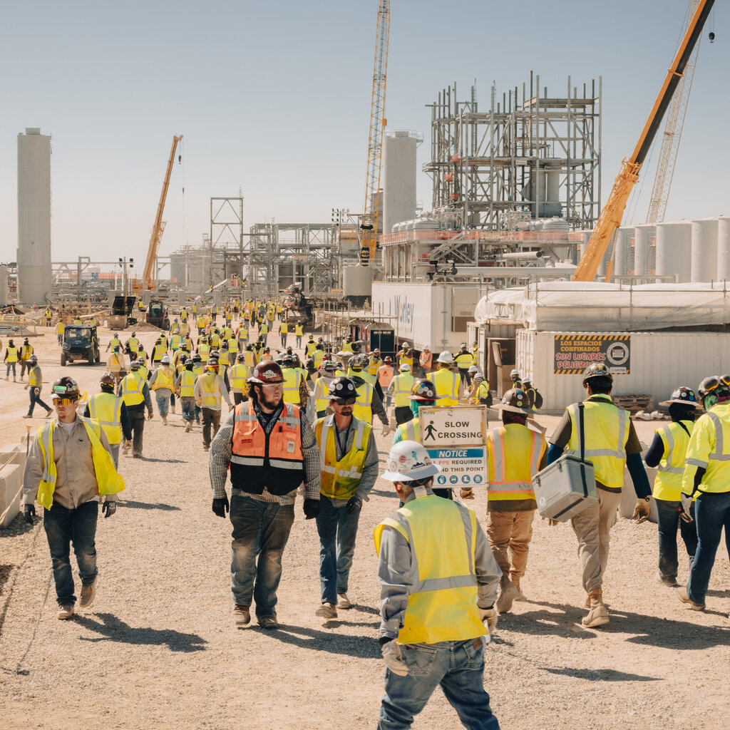 Dozens of people wearing yellow work vests, along with one person wearing bright orange, walk alongside an industrial construction site with numerous cranes and steel structures.