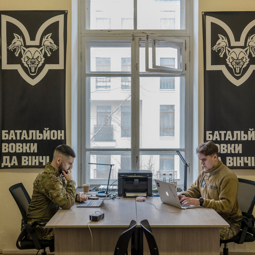 Two men in military uniforms sit at two desks facing each other, both working on laptops. 