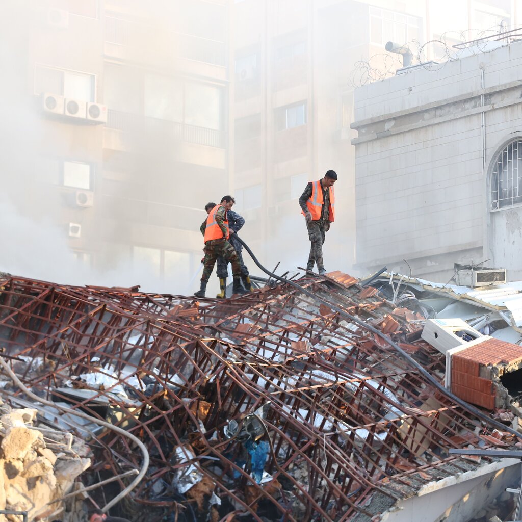 Three rescue workers stand atop a pile of rubble as smoke rises around them.