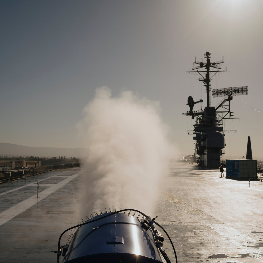 A long view down the flight deck of an aircraft carrier from the stern. The gray superstructure, with various masts and radar domes, is visible on the right. In the foreground, a device that looks lake a big fan is spraying a white mist. 