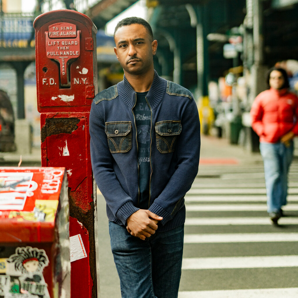 A man wearing blue jeans and a blue sweater leans against a red fire alarm pull station in New York. 