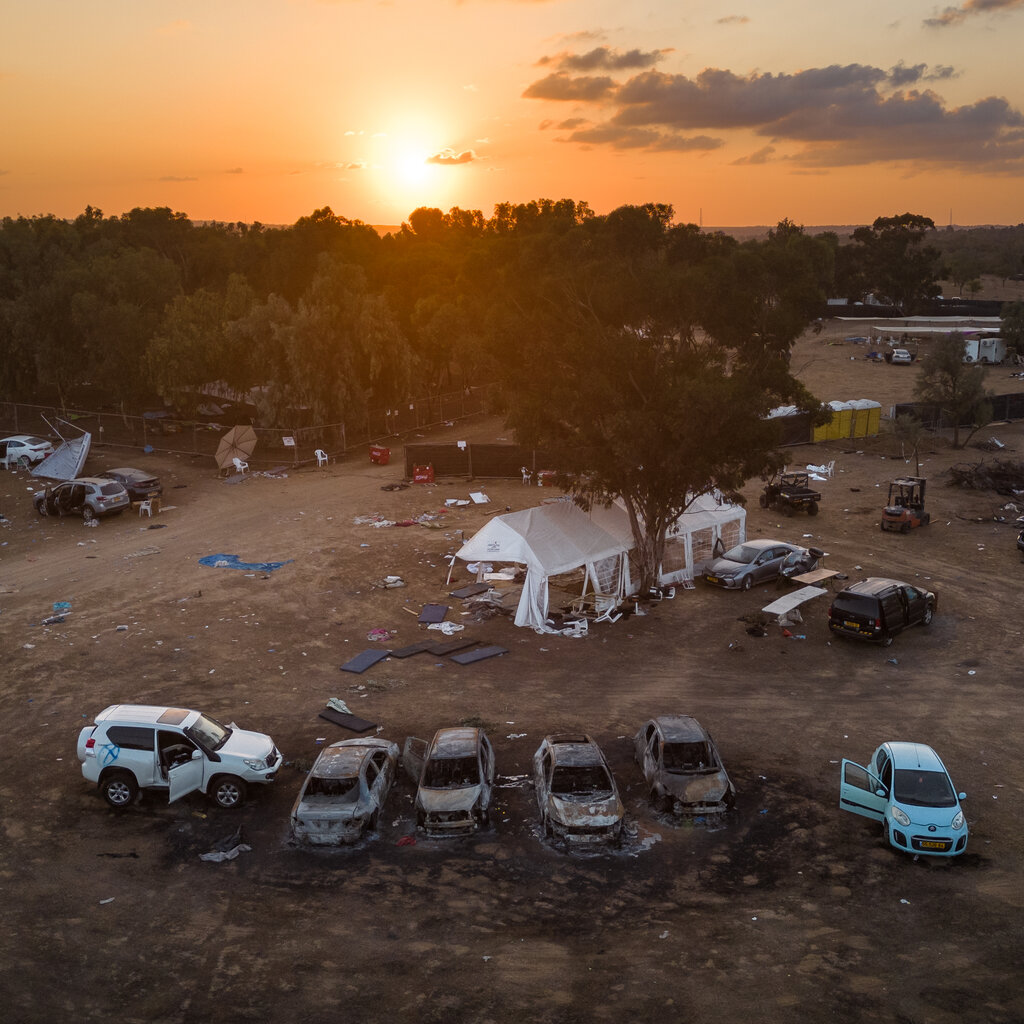 An aerial view of campground, with burned-out vehicles and abandoned tents, as the sun sets behind trees.