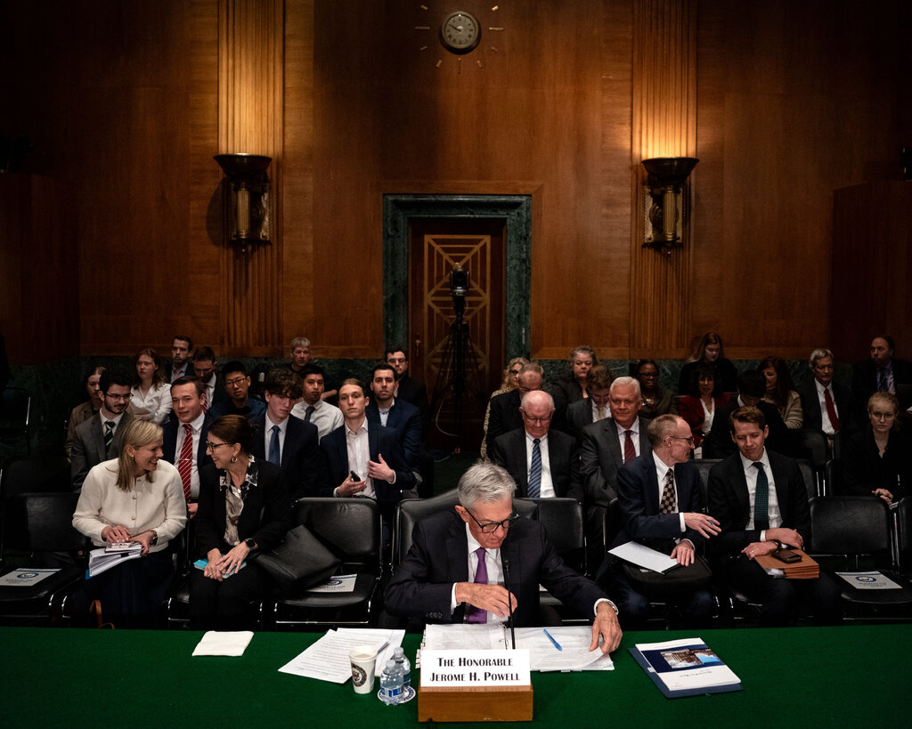 Jerome Powell sits at a desk in a Senate committee room, people are seated behind him to watch the testimony. 