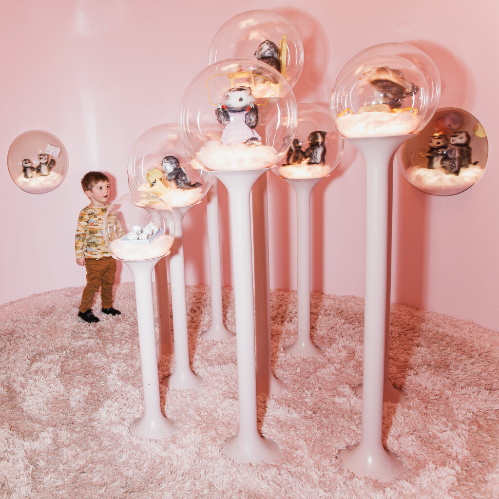 In this photograph, a small boy in brown pants enters a small house with pink walls and a pink rug. In the room are seven bubbles atop pink pedestals, each containing characters from the Frances books by Russell and Lillian Hoban.
