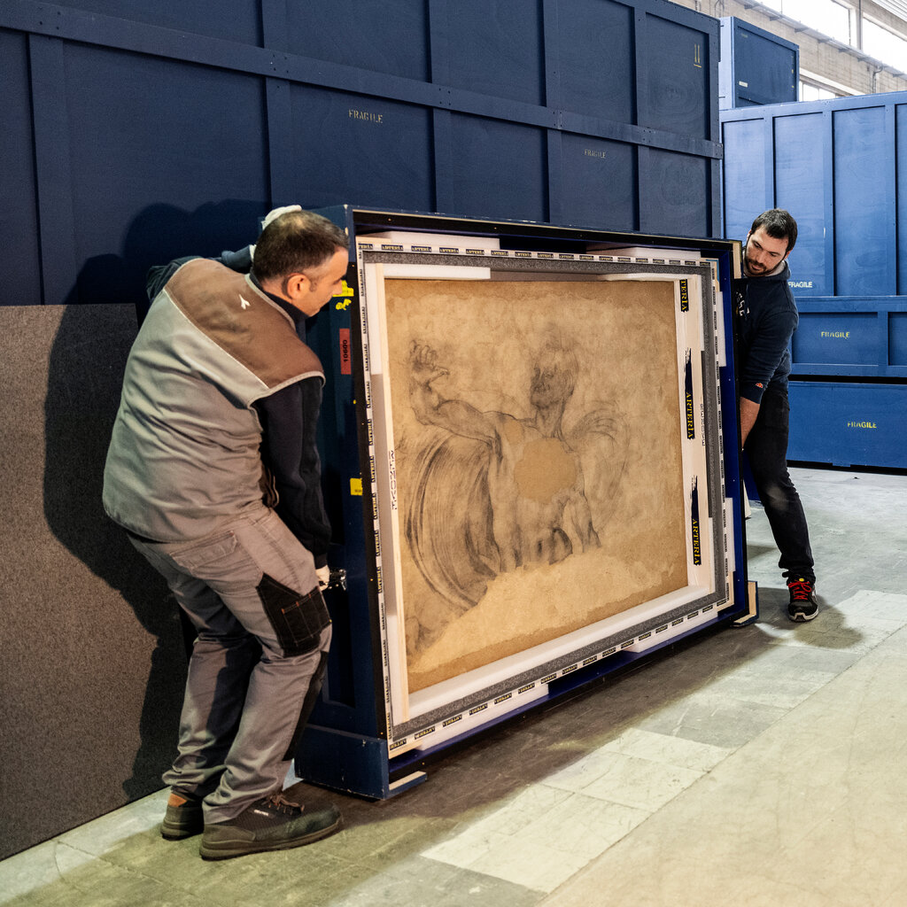 In a warehouse, two men handle a large framed drawing.