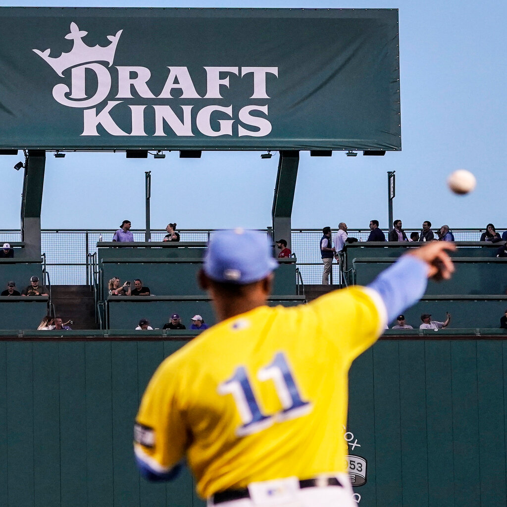 A baseball player in a yellow and blue jersey throws a baseball in front of a billboard that reads "Draft Kings."
