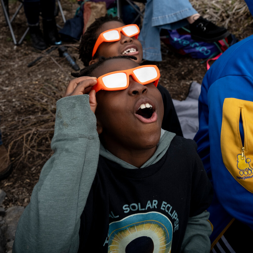 Two children, wearing orange glasses, look up in the sky. The boy in the foreground opens his mouth in wonder. 