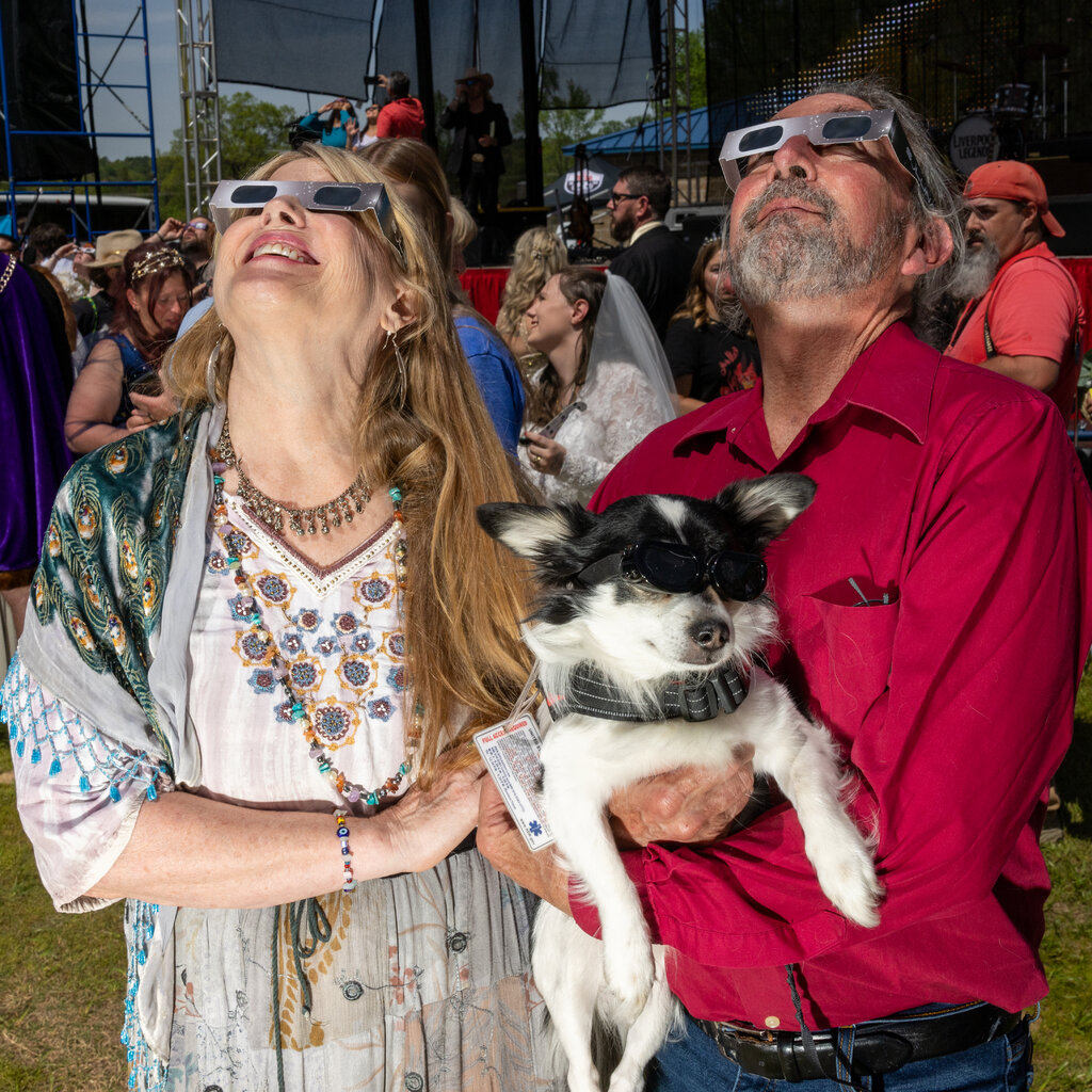 A woman and a man stare up at the sky wearing eclipse glasses. The man, wearing a deep red button down shirt, is holding a dog. The dog also has glasses on. 