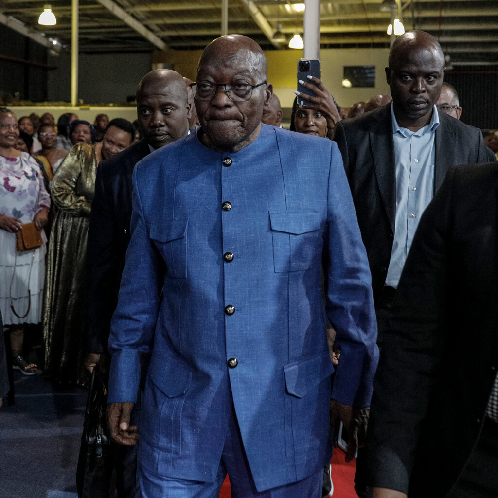 Jacob Zuma, in a bright blue suit, walking at an event in a big room with a lot of people.
