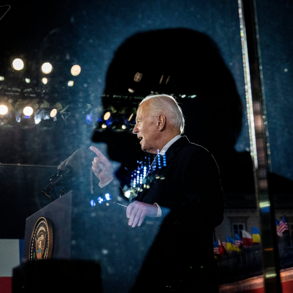 President Biden speaking at a lectern outdoors at night, seen through a window.