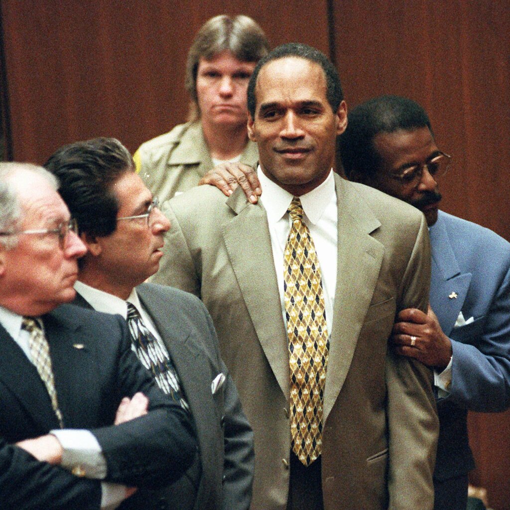 O.J. Simpson wearing a tan suit and yellow patterned tie as he is embraced from behind by his lawyer, Johnnie Cochran.