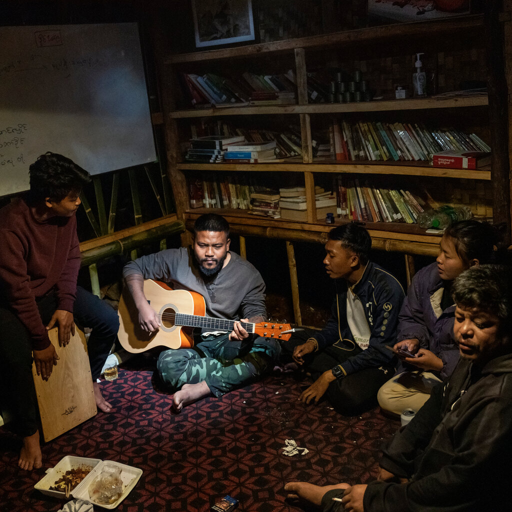 In a dimly lit room with books on shelves, a bearded man sits on the floor playing an acoustic guitar, as five people sit around him.