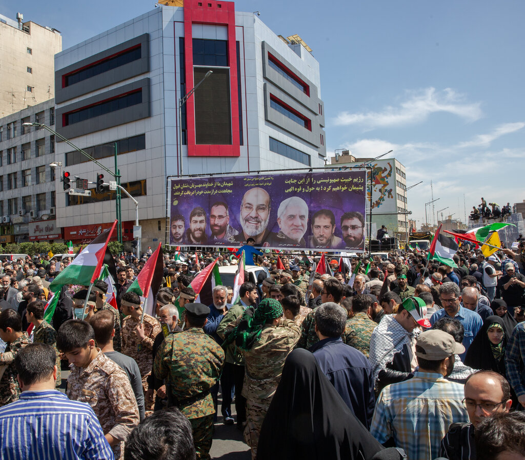 A giant poster with images of seven men looms above a dense crowd carrying flags on the street during the day.