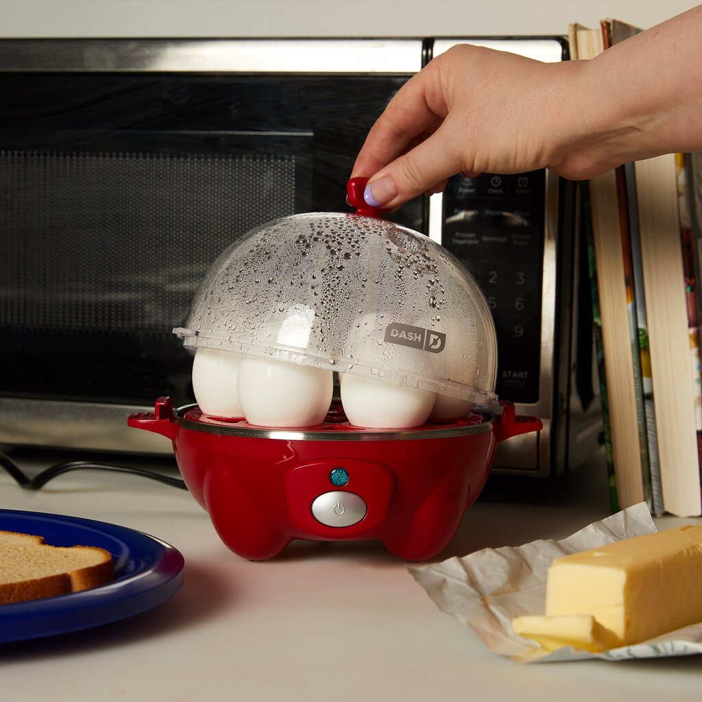A hand lifting the lid of a red Dash Rapid Egg Cooker with six eggs in it. 