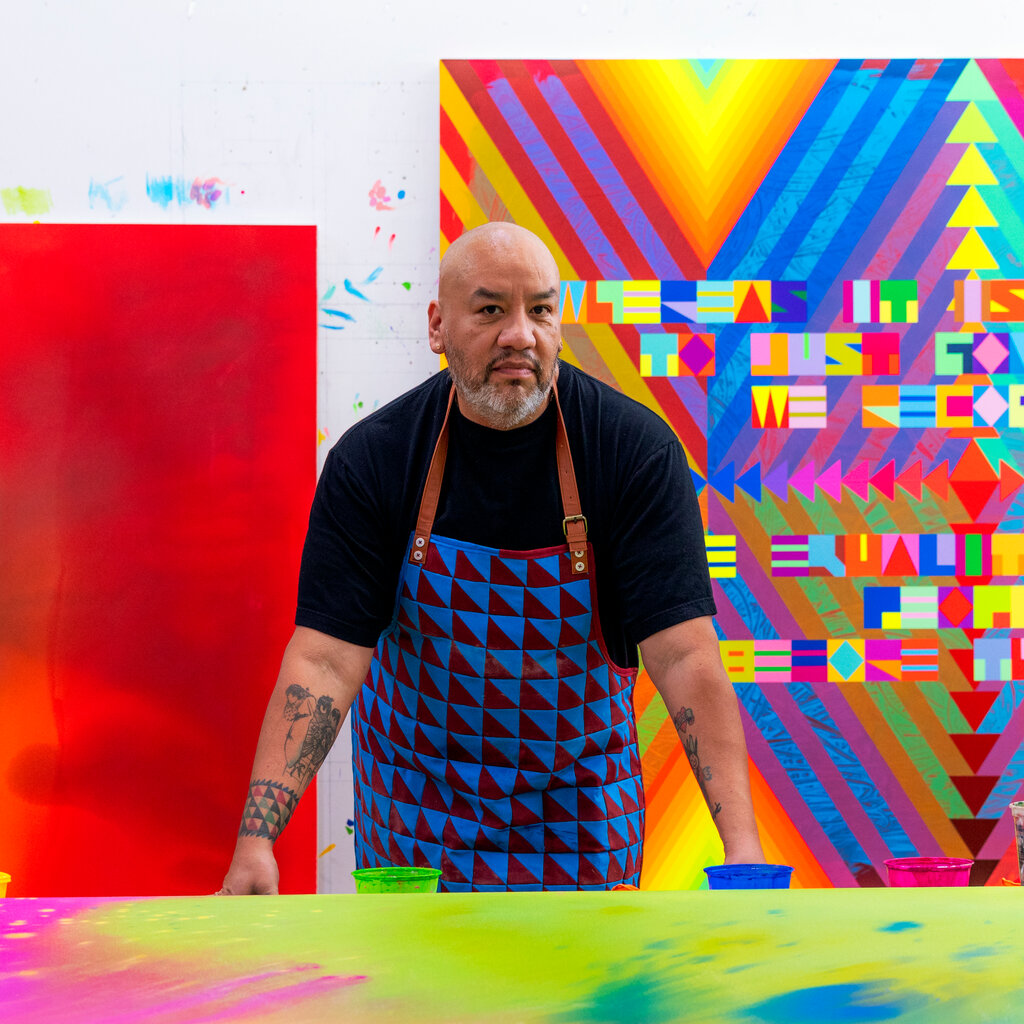 A man in a blue apron with red triangles stands in front of a swirling psychedelic painting he is in the process of creating in blue, pink and chartreuse. At left is a crimson and blue painting and at right a painting of eye-dazzling crosses with inset Native symbols.