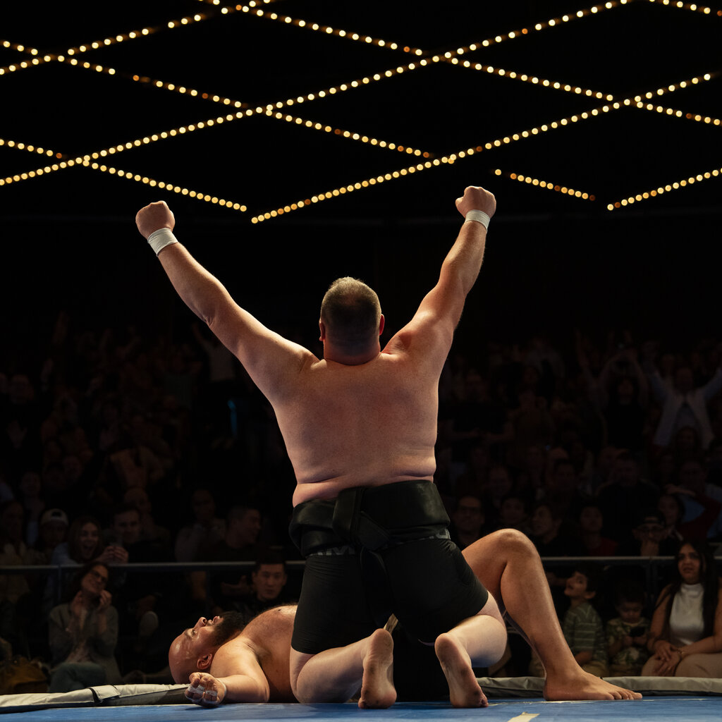 A sumo wrestler, kneeling, raises his arms in victory in front of another wrestler lying face up on the mat. 