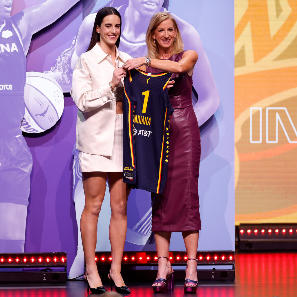 Caitlin Clark in a white outfit, stands next to the WNBA commissioner. Both are holding a single Indiana jersey. 