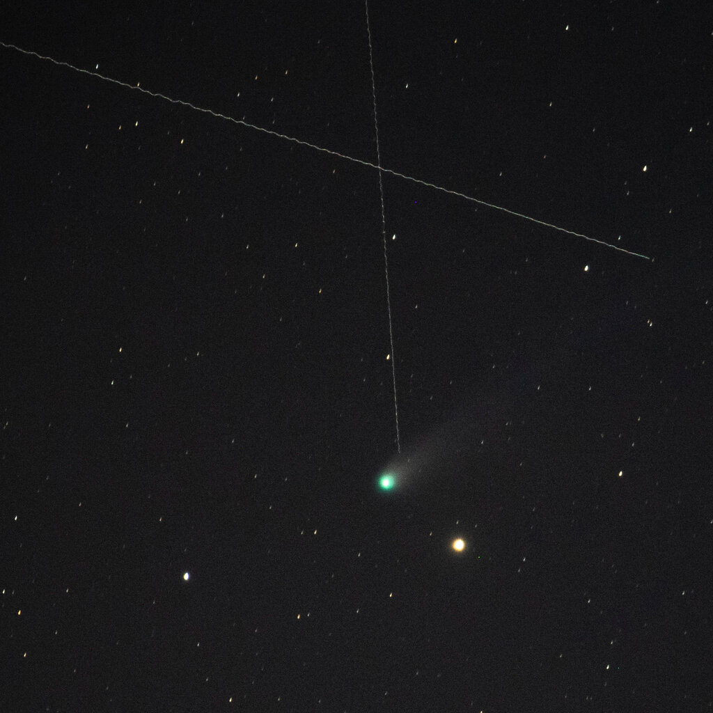 A greenish comet with its tail in the night sky passes a yellow star, with two streaks from satellites above them photographed during a long exposure.