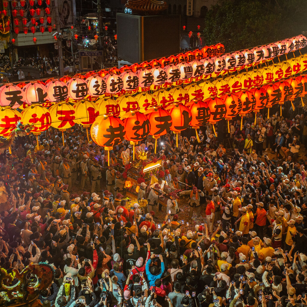 A sea of people look and take images of a procession of a sedan chair holding a goddess. Red lanterns are above them. 