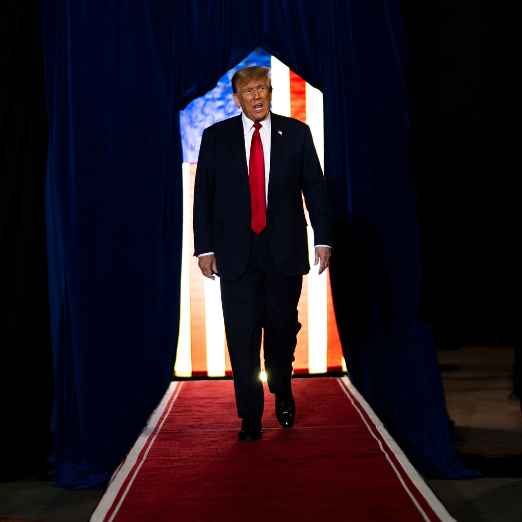 Donald Trump walks a red carpet at a rally, his mouth open.