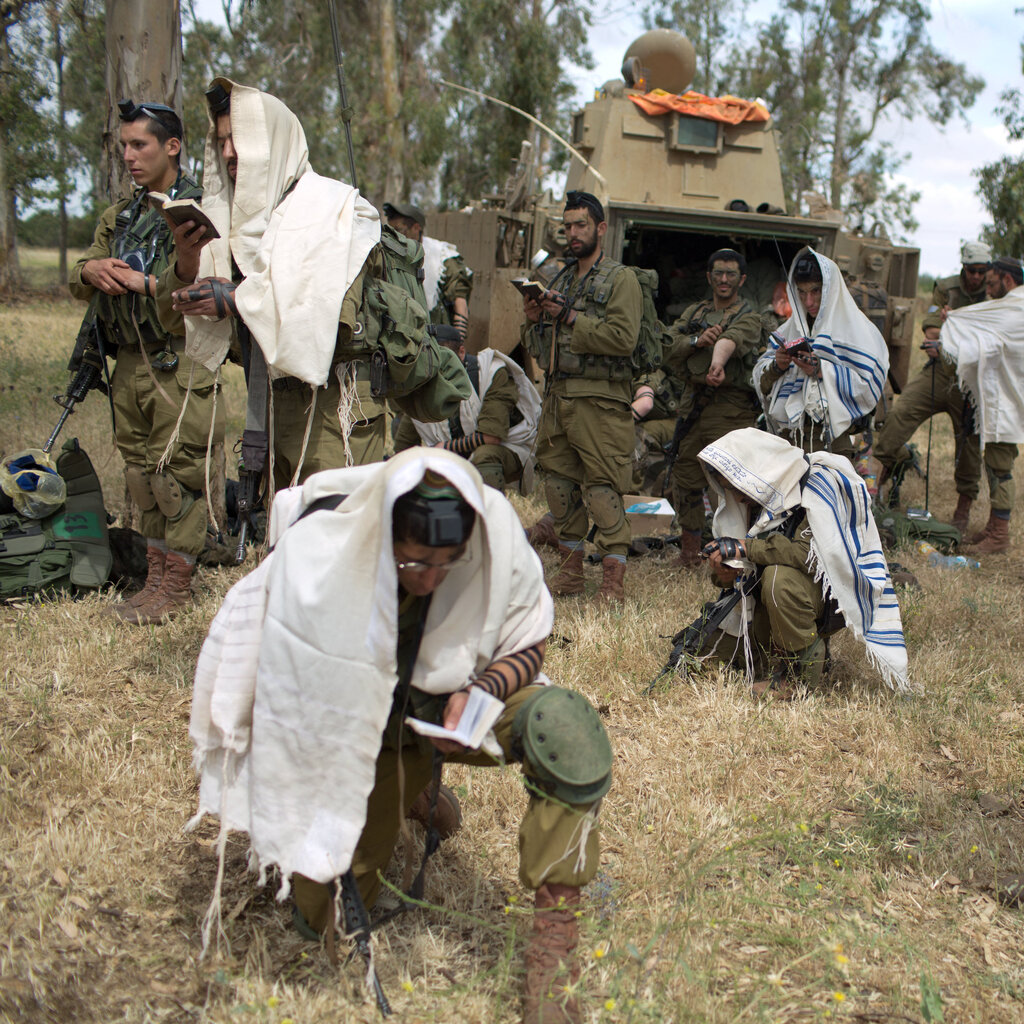 Soldiers in fatigues, with some wrapped in fringed prayer shawls. One is kneeling and looking at a prayer book.