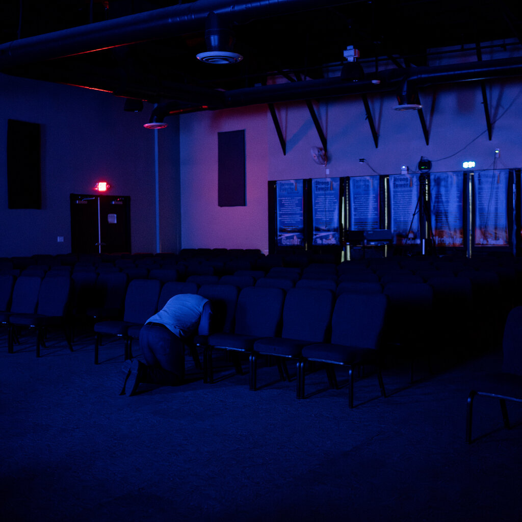 Camilo Perez seen from behind praying on his knees in a blue-lit room.