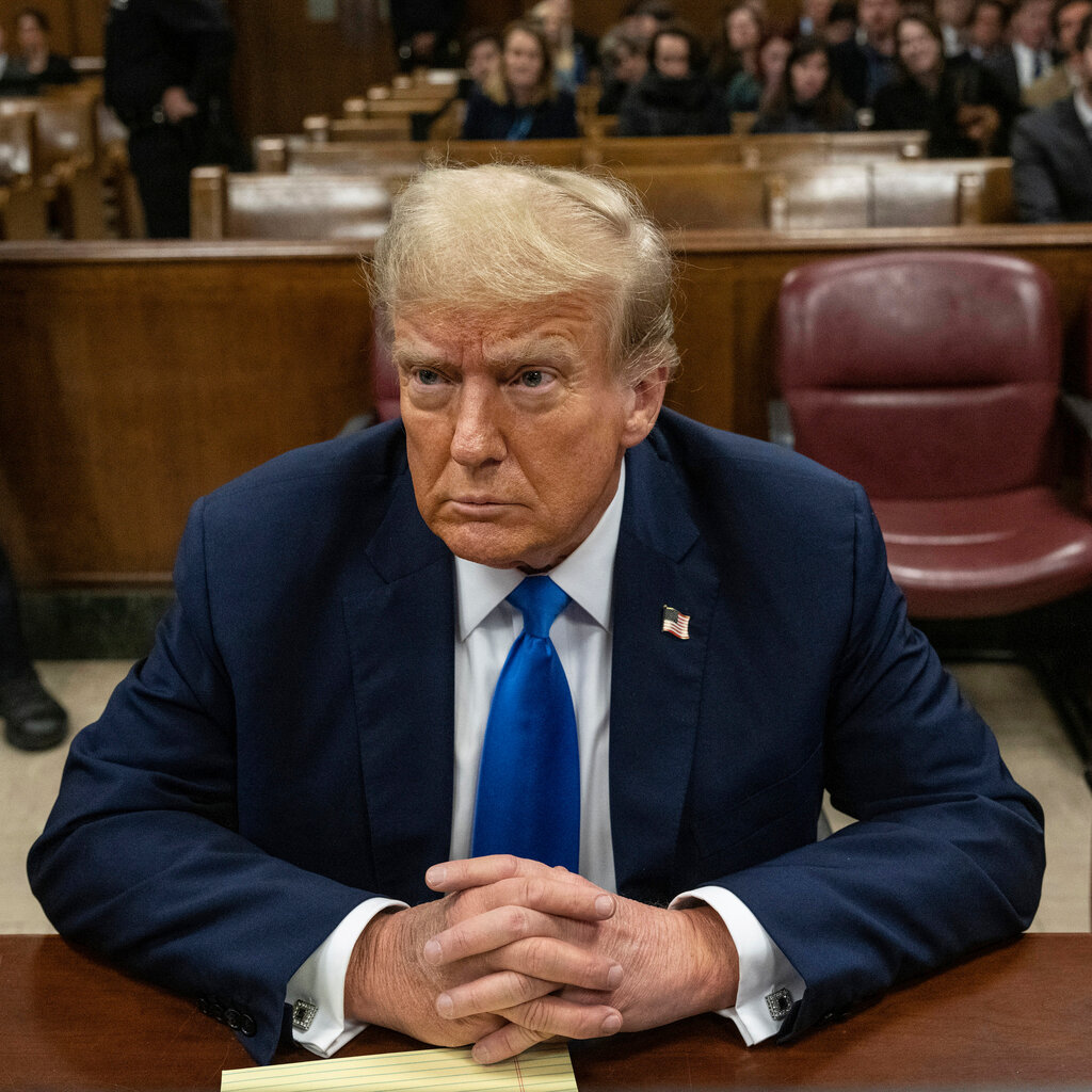 Donald Trump sits, unsmiling, at a desk in a courtroom. 