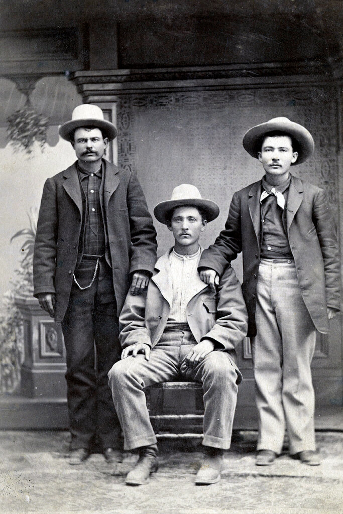 In a black-and-white portrait, three young men pose in ranch-hand attire: jackets, heavy trousers, Western hats and boots.
