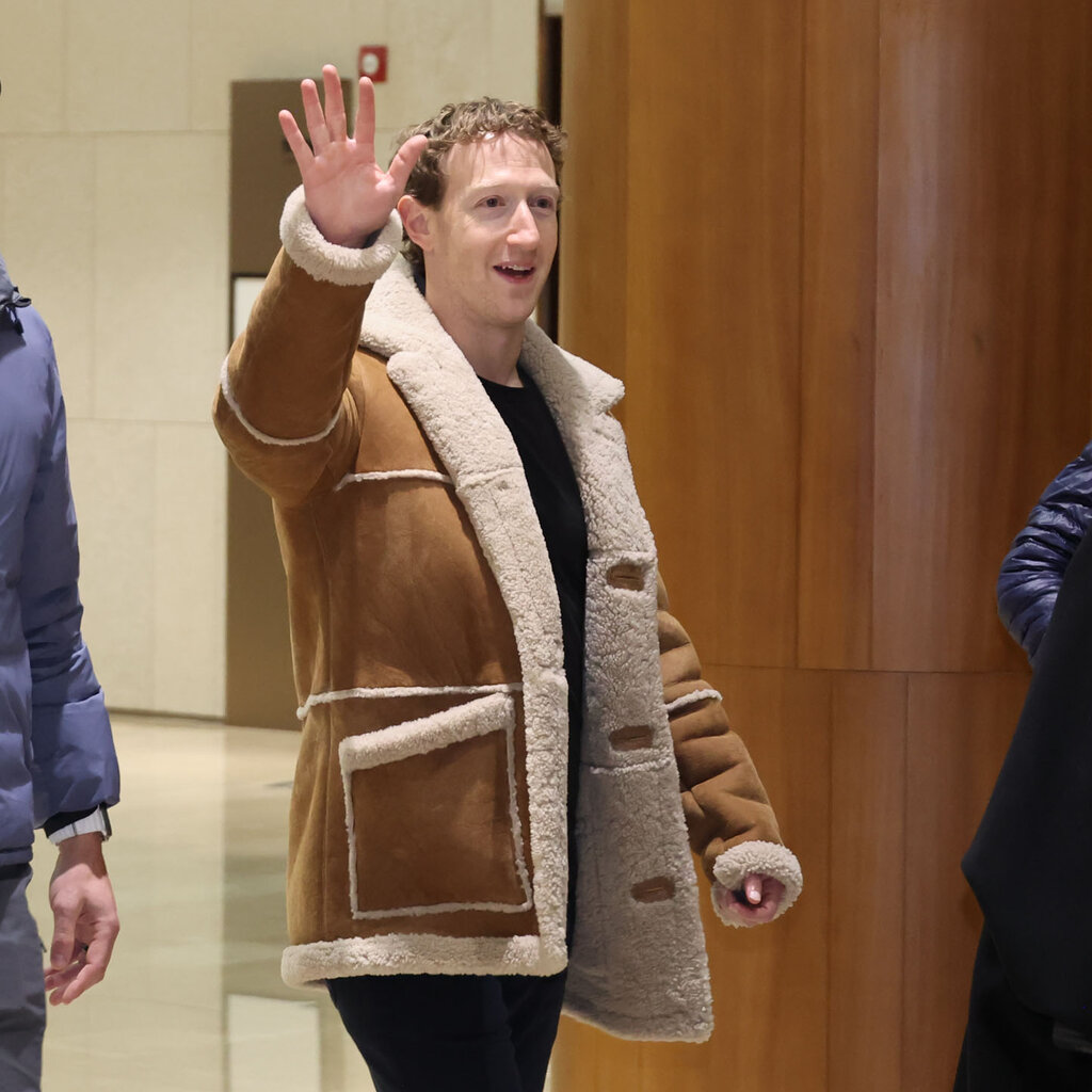 Mark Zuckerberg, wearing a tan shearling jacket, waves toward photographers. 