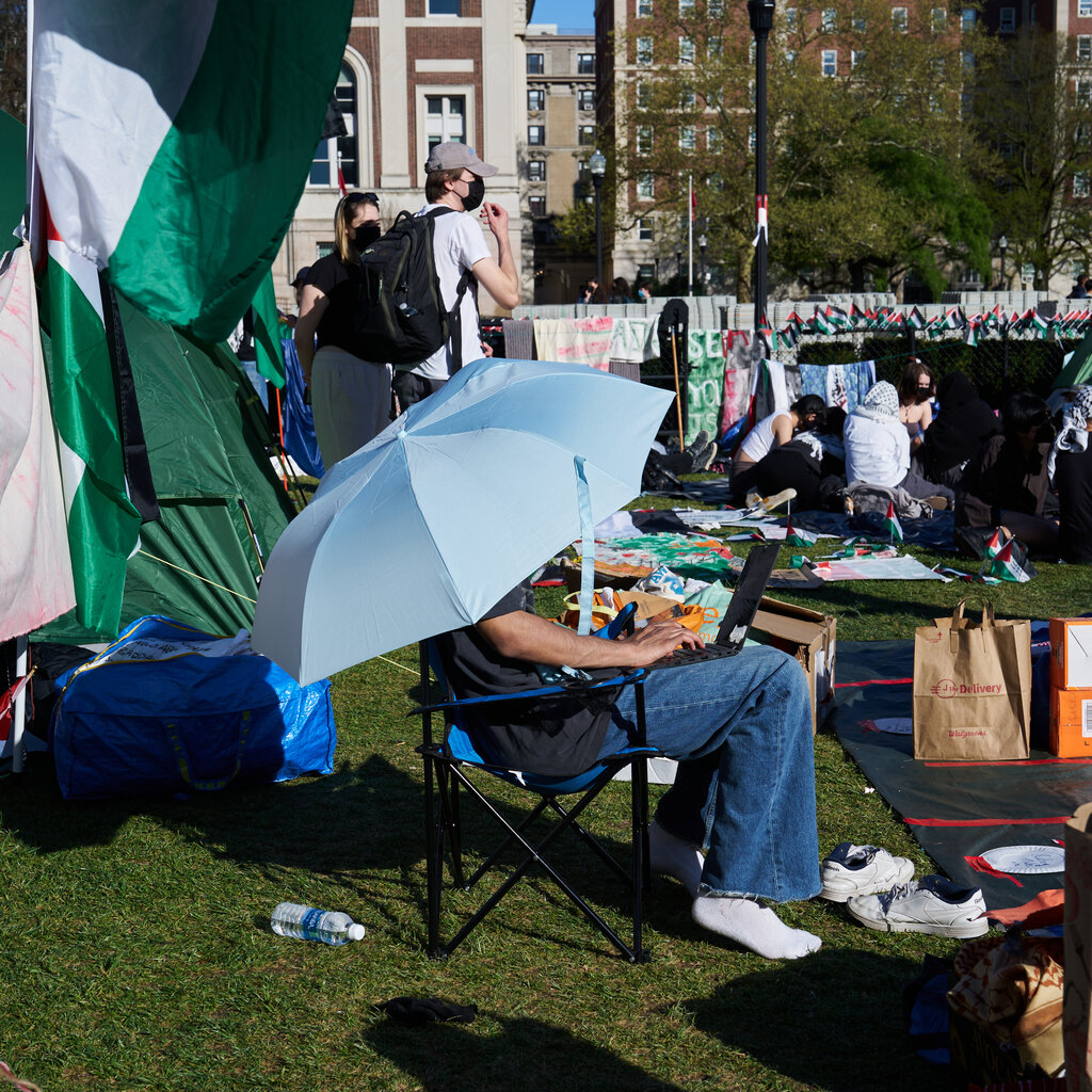 A student sits within a protest encampment under the shade of an umbrella, working on a laptop. 