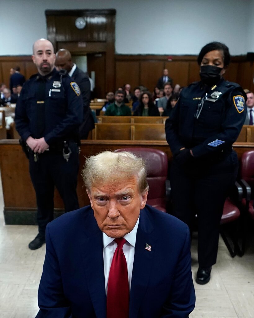 Donald Trump sits in a courtroom; two police officers stand either side behind him. 