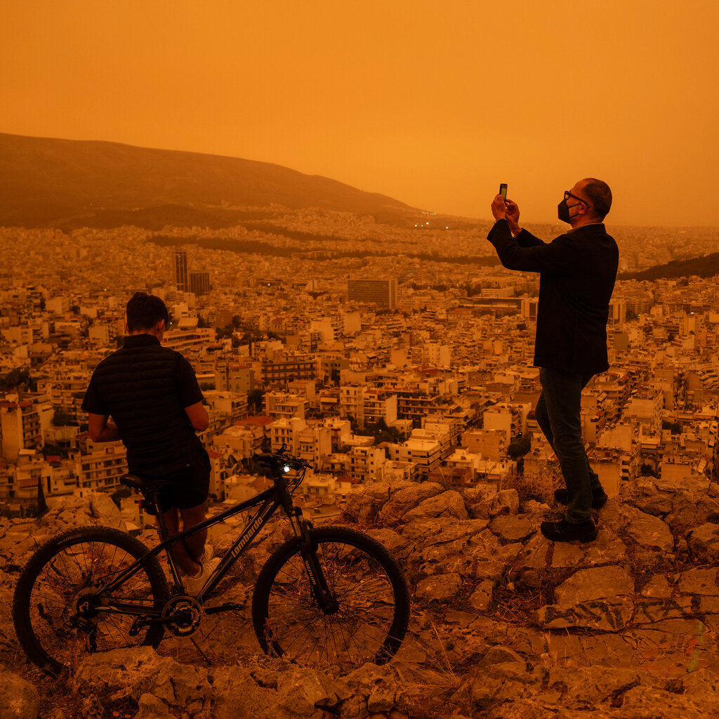 A man on a hill wearing a face mask holds a camera and takes a picture of Athens under an orange sky, while a man to his left leans on a bicycle. 