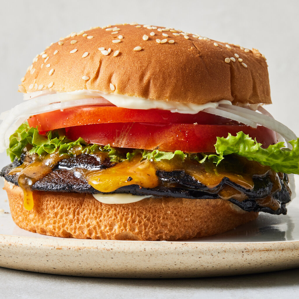 A mushroom smash burger with lettuce, tomato, onion and melted cheese on a sesame seed bun sits on a beige plate.