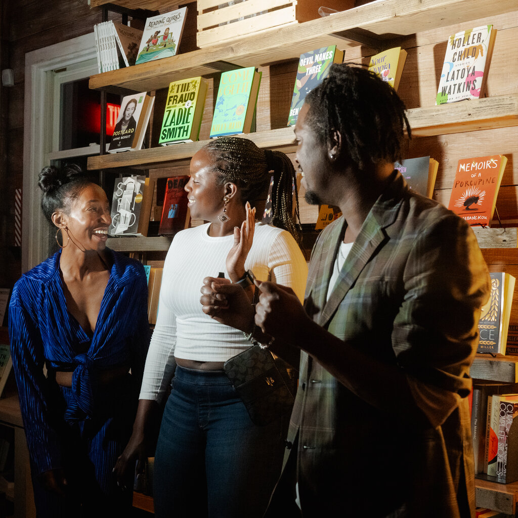 Three people stand, talking, against a book shelf, 