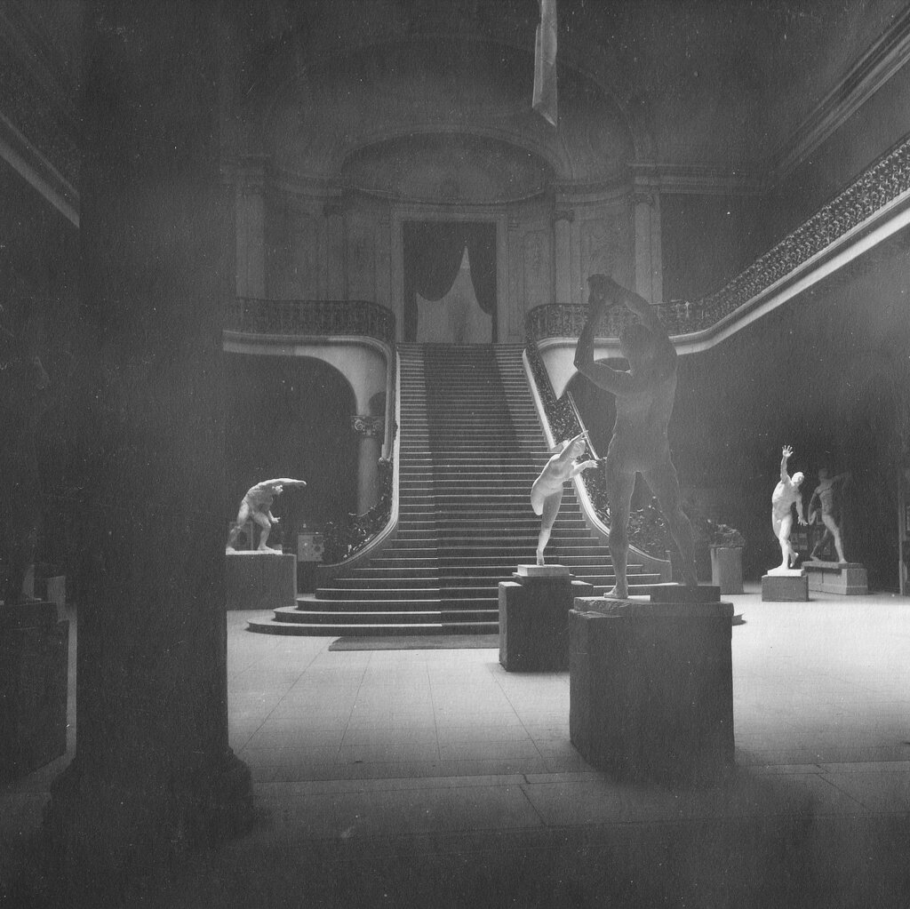 A black-and-white image of statues in a cavernous room with a grand staircase. 