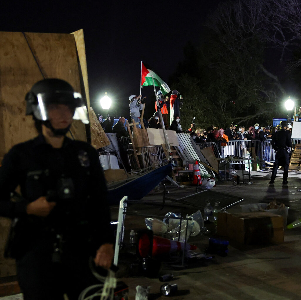 Demonstrators behind makeshift barricades of wood and metal barriers hold a Palestinian flag aloft. Two security officers are on guard.