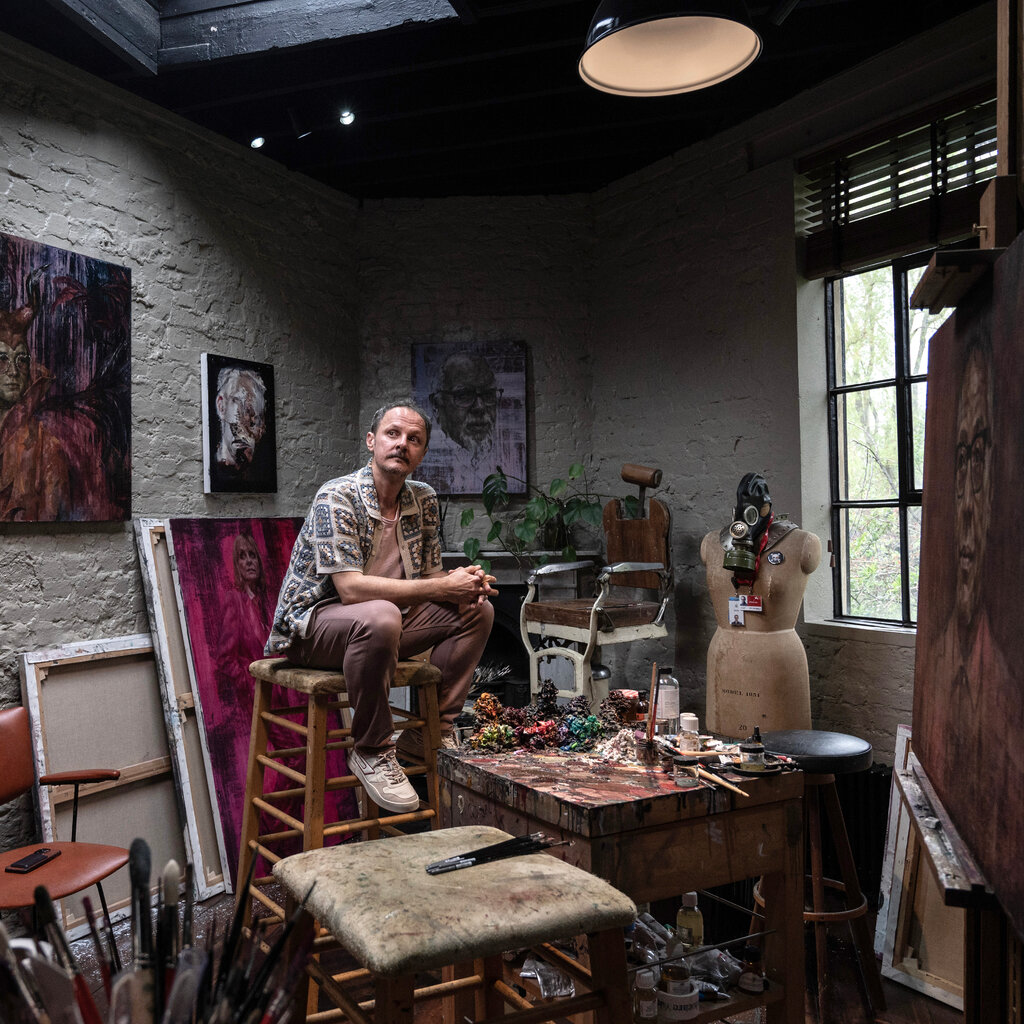 Jonathan Yeo sits on a tall stool in a workshop with pictures on the whitewashed walls and brushes and other painting paraphernalia scattered around.