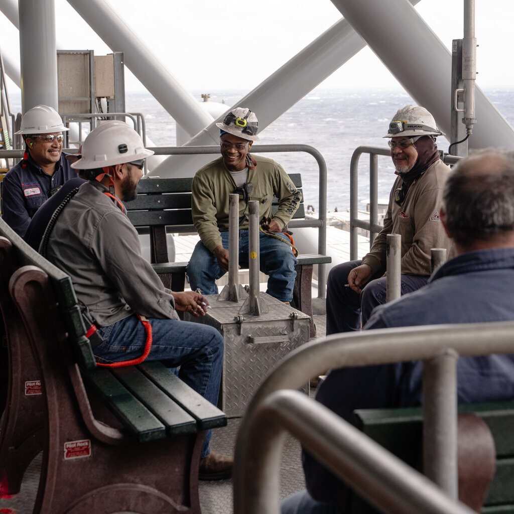 Workers in hard hats sit on an oil platform and smile at each other. The ocean is visible behind them. 