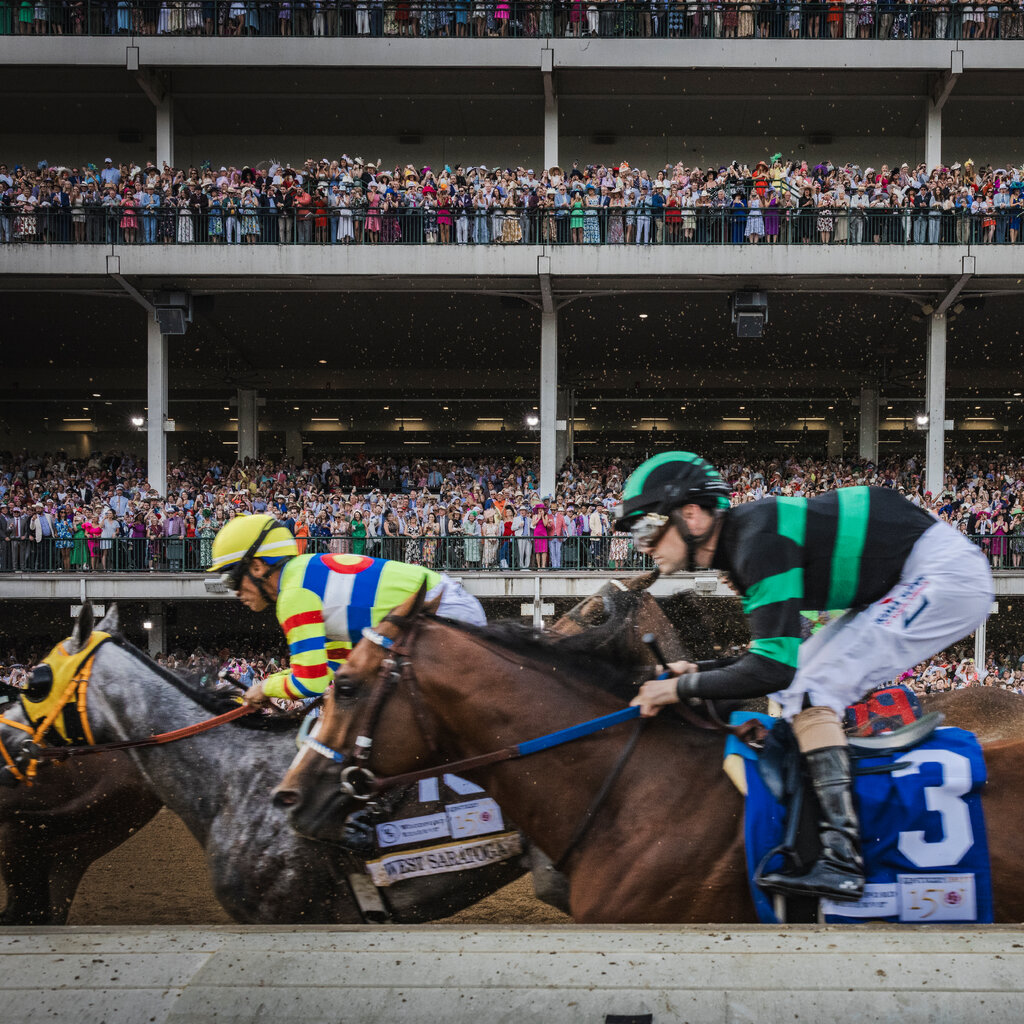 In the foreground, a jockey in black and green clothing rides a horse wearing the No. 3. A second horse, ridden by a jockey in multicolored clothing, is behind them. Crowded viewing stands are in the background.