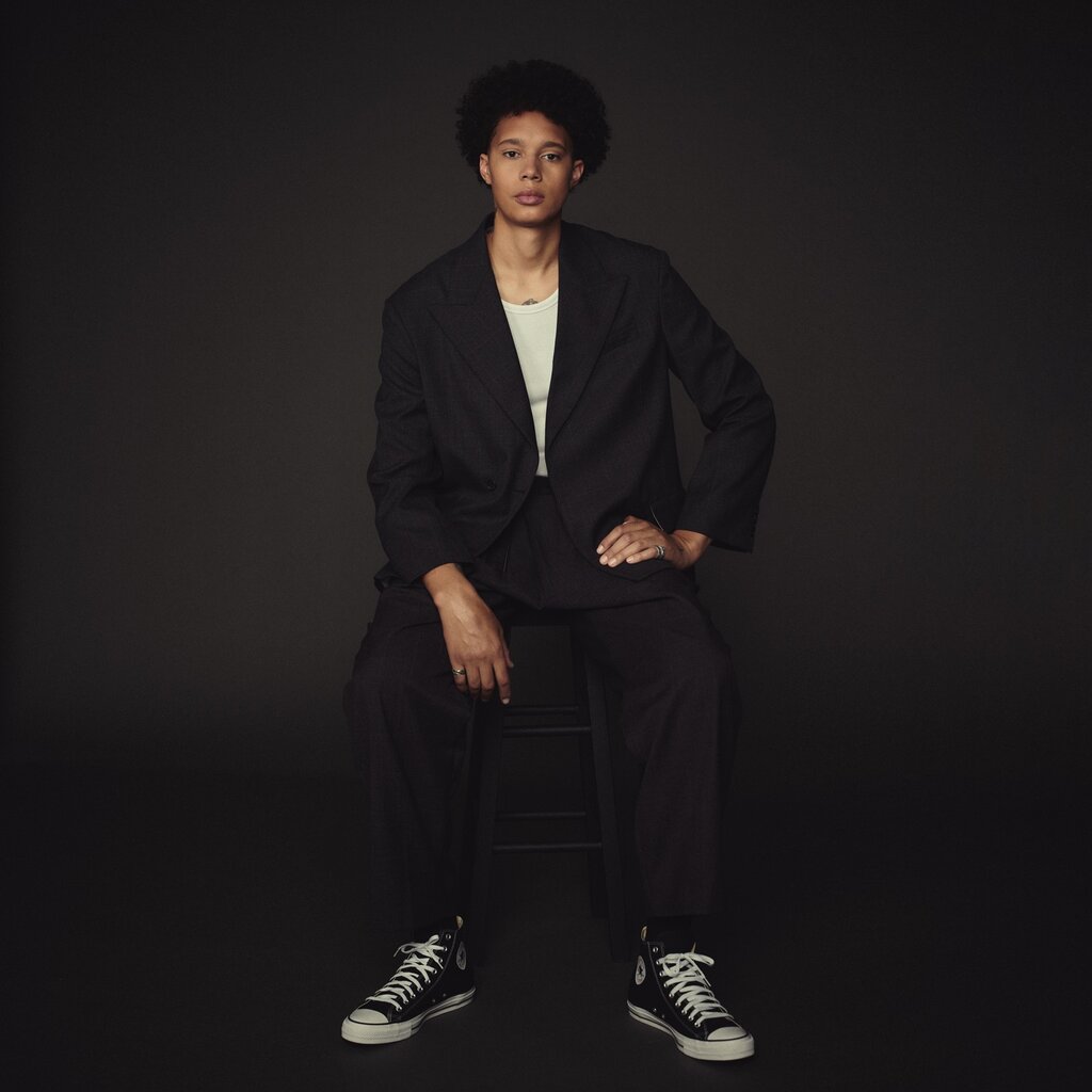 A portrait of Brittney Griner in a dark suit and sneakers sitting on a stool.