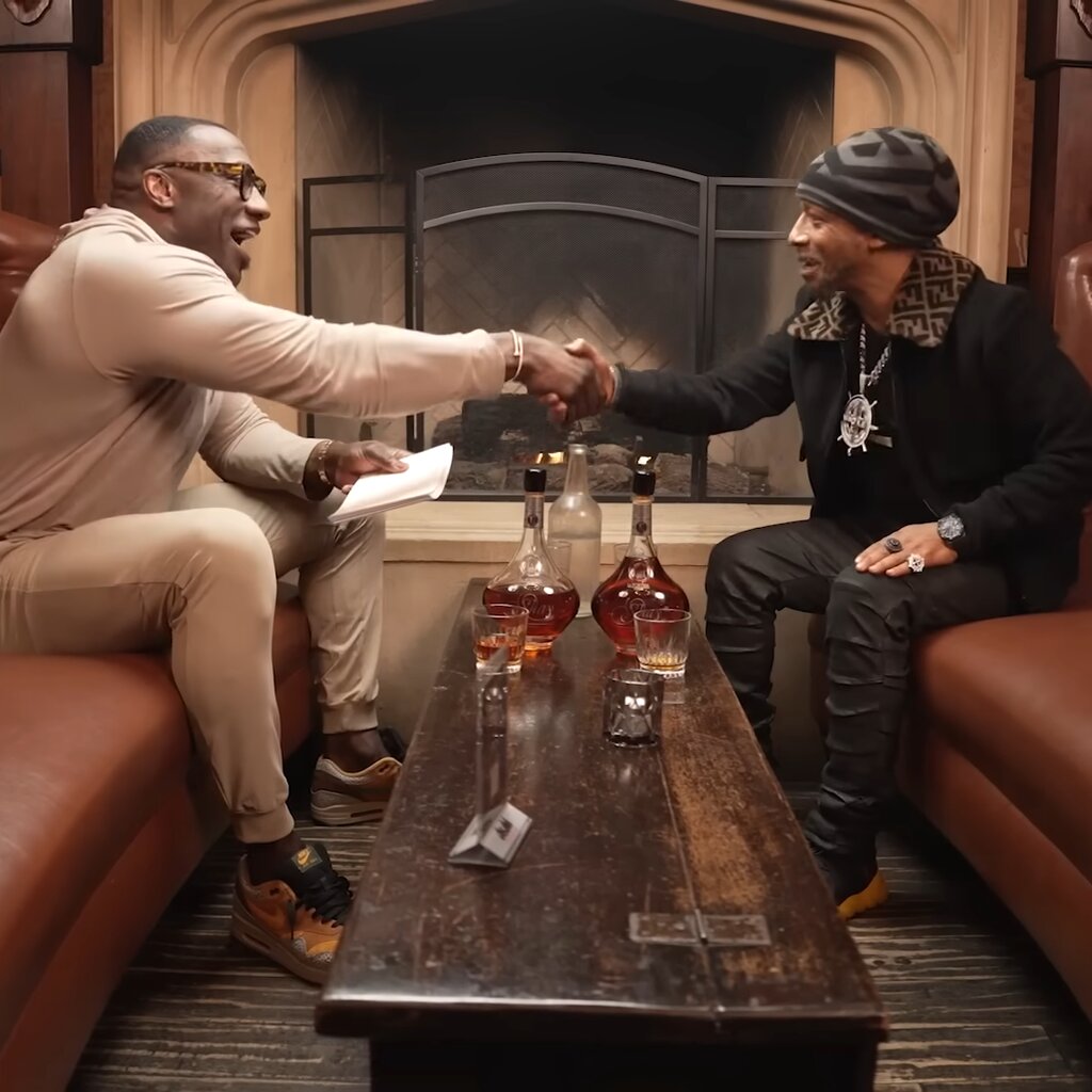 Shannon Sharpe shakes the hand of the comedian Katt Williams over a coffee table holding bottles of cognac.