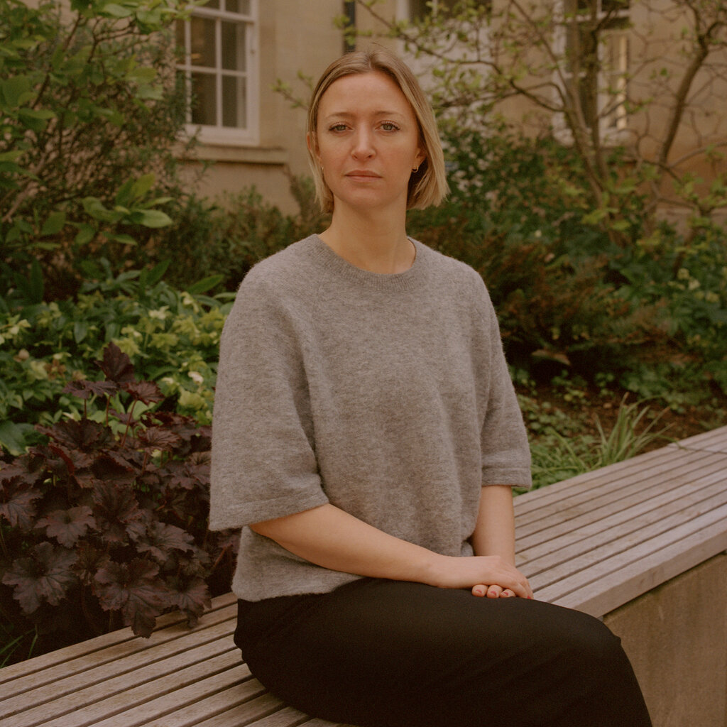A portrait of Lucy Foulkes, who wears a gray sweater and black pants and sits at a garden area outside the department of experimental psychology in Oxford, England.