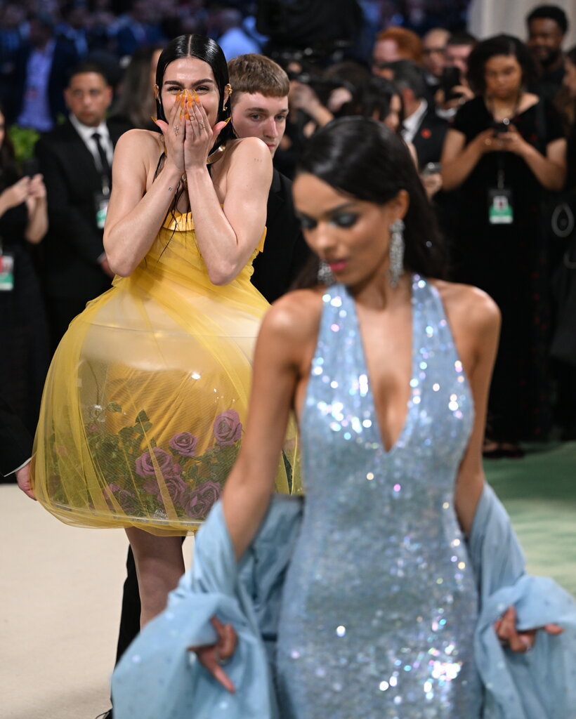 A woman in the background wears a yellow translucent dress that contains a terrarium. A woman in the foreground wears a baby-blue sequined gown with a shawl.