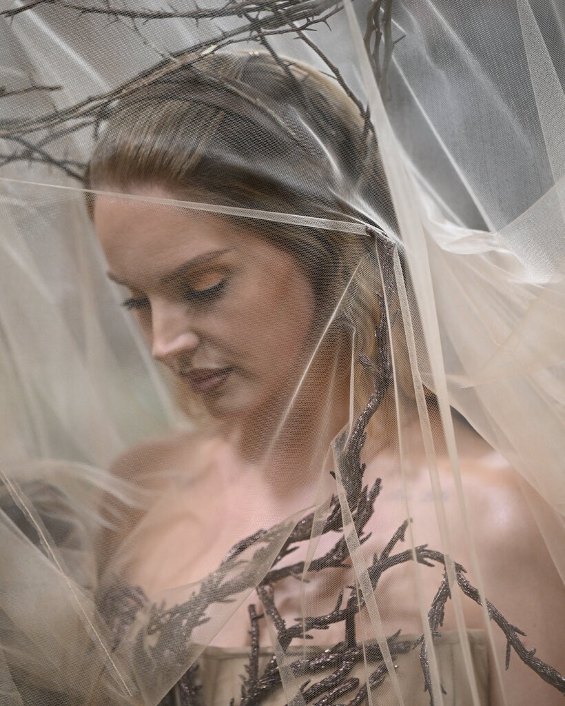 A woman wears a translucent veil over tree branches that encircle her body.