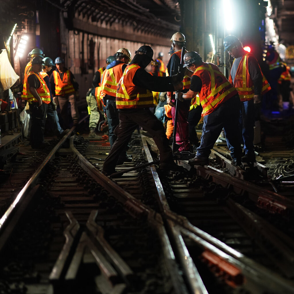 M.T.A. workers wearing hard hats and orange work vests repair subway tracks. 