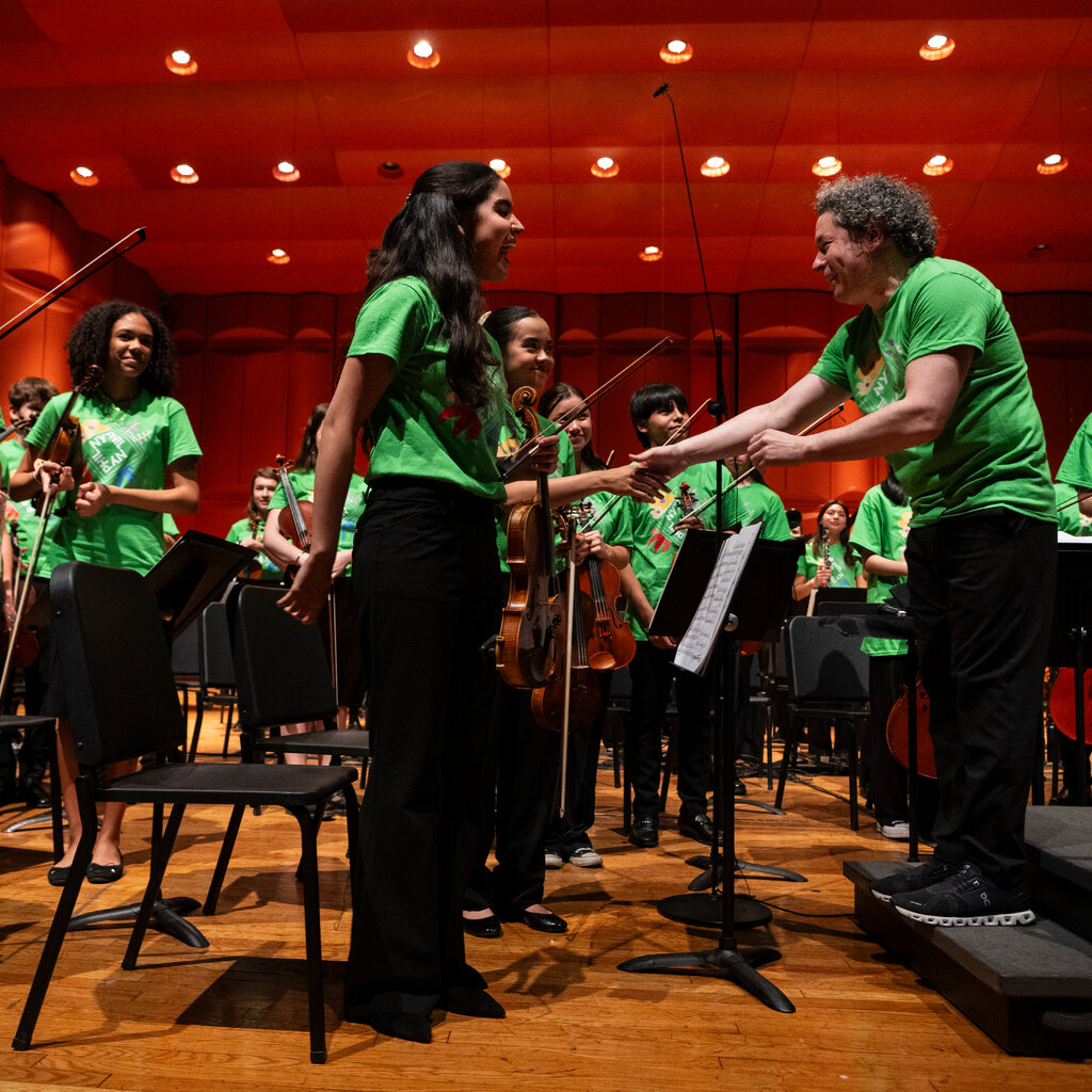 Gustavo Dudamel shakes hands with a young violinist as other young musicians in the orchestra look on. Everyone is wearing a green T-shirt. 