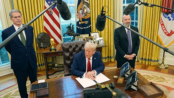 Donald Trump sits behind the desk in the oval office surrounded my microphones as cabinet members look on.