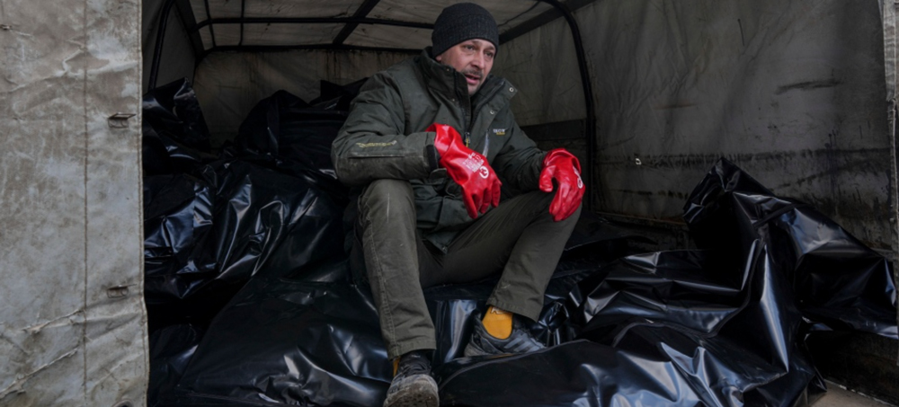 A mortuary worker sits on body bags before they were transported to be buried in a mass grave on the outskirts of Mariupol, Ukraine, Wednesday, March 9, 2022. (photo: Evgenly Malotetka/AP)