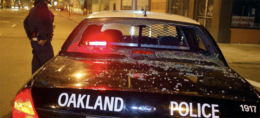 An Oakland Police officer stands next to his car with the rear window broken out January 26, 2003 in Oakland, California after the Oakland Raiders lost to the Tampa Bay Buccaneers in Super Bowl XXXVII. (photo: Justin Sullivan/Getty Images)